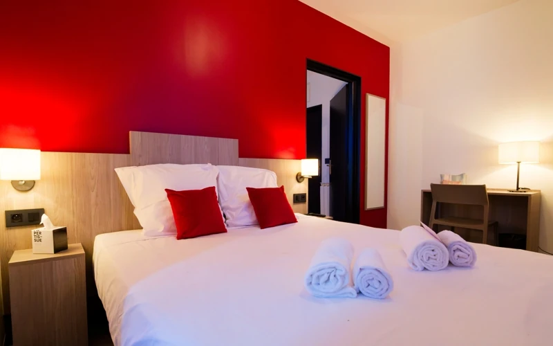 Vue d'ensemble du lit double avec mur rouge théâtral, chevets et coin bureau à l'Hôtel des Artistes